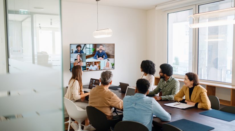 In a well-lit meeting room, a team actively engages with remote colleagues via a large screen, illustrating the modern hybrid work model where technology bridges the gap between in-office and remote team members.