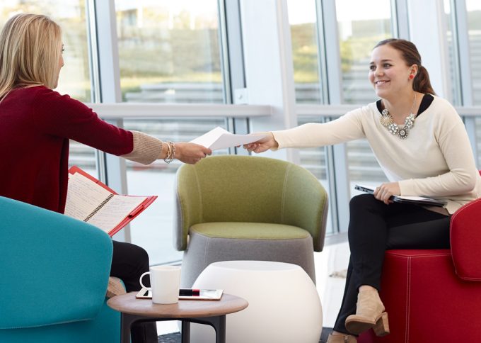 A woman extending a piece of paper to another woman who is reaching for it.